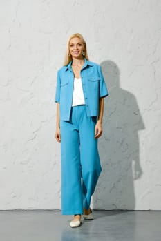 Smiling woman in her fashionable spring outfit standing against a wall, blond hair and blue eyes looking into the camera.