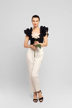 Beautiful adult woman elegantly posed with flower in hand in a black ruffled blouse and white trousers, looking straight at the camera in an indoor studio shot.