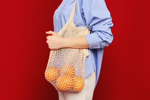 Unrecognizable woman holding oranges in a string bag on ruby red background. Concept of sustainable living.