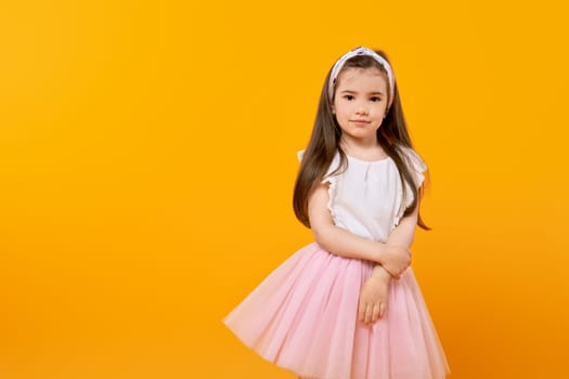 Cute little girl stands in a yellow-tinted studio, looking into the camera with innocence. Her long hair and fashionably adorned dress reflect her childhood as she captures this moment of beauty.