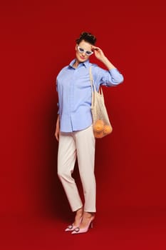 Trendy woman with oranges in reusable string bag over her shoulder touching her sunglasses on ruby red background. Concept of sustainable living and care of the environment.