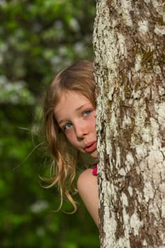 A blonde girl looks out from behind a tree