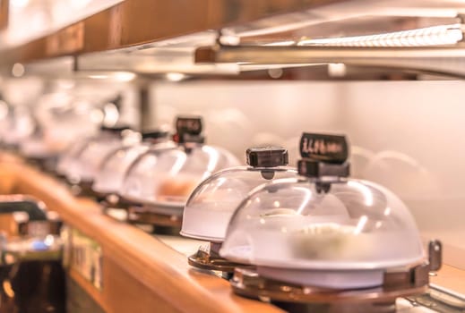 Close-up on Japanese Conveyor belt sushi restaurant where plates with sushi are placed on a rotating rail protected with a clear plastic lid.