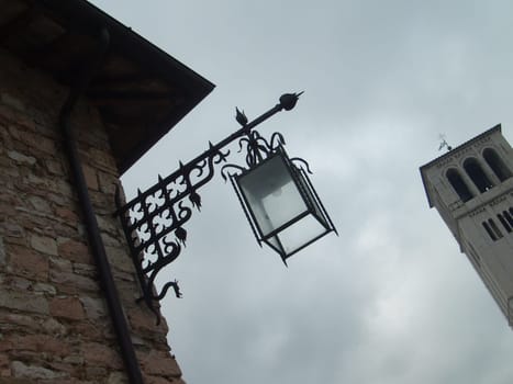 Assisi, Italy, December 1, 2007 Photo taken from an angle. Bottom up view of an old Italian tower and a beautiful metal street lamp. Brick wall of a building with an arrow-shaped lantern.