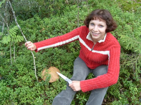 The girl found a big beautiful mushroom. A woman rejoices in a mushroom found in the grass and moss. She is wearing a red sweater with a zipper. The face is cheerful and surprised. In hand a knife.