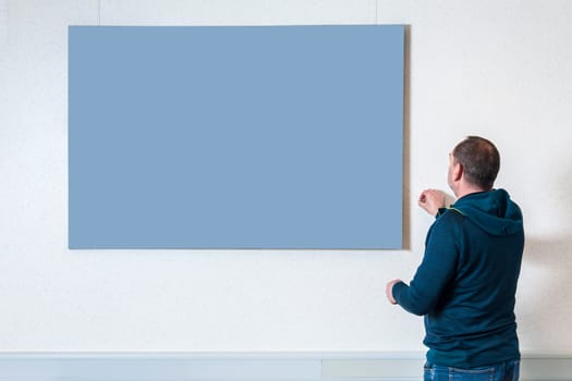 Upper body view of a man in casual clothing with his back to the camera adjusting a neutral image on a light colored wall.