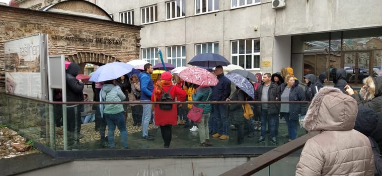 Sarajevo, Bosnia and Herzegovina, March 8, 2020. A group of tourists listening to the guide. It is raining, people are standing under various umbrellas. Ancient buildings made of stone. Bezistan.