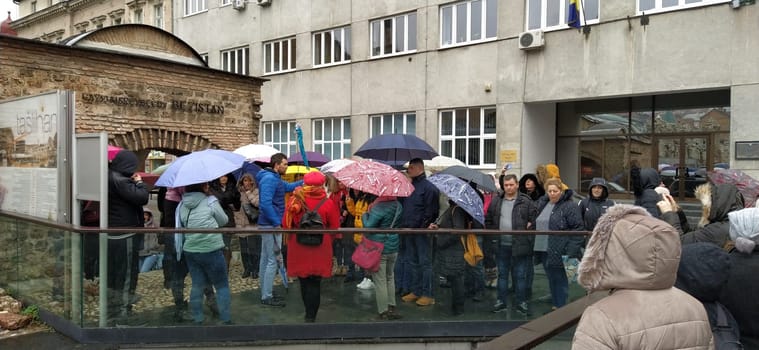 Sarajevo, Bosnia and Herzegovina, March 8, 2020. A group of tourists listening to the guide. It is raining, people are standing under various umbrellas. Ancient buildings made of stone. Bezistan.