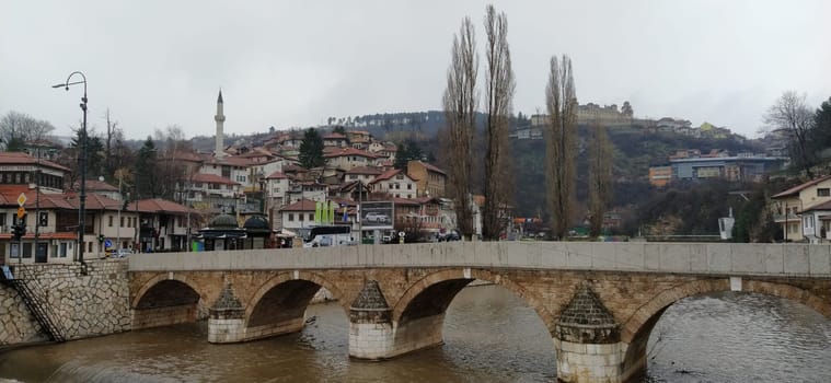 Sarajevo, Bosnia and Herzegovina - March 8, 2020: Sheher bridge over the Milatsk river. Spring. Turbid water in the river and bare trees. Turkish architecture and mosque. Mountains on the horizon.