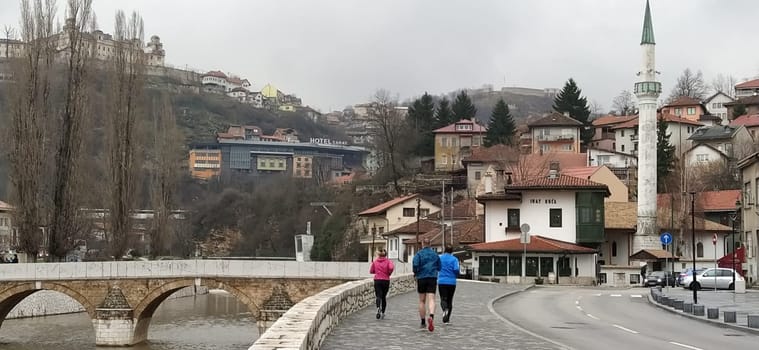 Sarajevo, Bosnia and Herzegovina - March 8, 2020: Sheher bridge over the Milatsk river. Spring. Turbid water in the river and bare trees. Turkish architecture and mosque. Mountains on the horizon.