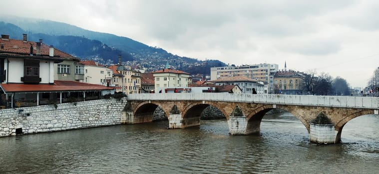 Sarajevo, Bosnia and Herzegovina - March 8, 2020: Sheher bridge over the Milatsk river. Spring. Turbid water in the river and bare trees. Turkish architecture and mosque. Mountains on the horizon.