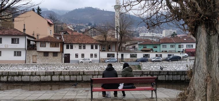 Sarajevo, Bosnia and Herzegovina - March 08, 2020. A young couple sitting on a wooden bench under a tree. The embankment of the river Milyack. View of the city of Sarajevo, a mosque and a minaret. Spring
