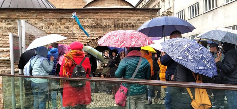 Sarajevo, Bosnia and Herzegovina, March 8, 2020. A group of tourists listening to the guide. It is raining, people are standing under various umbrellas. Ancient buildings made of stone. Bezistan.