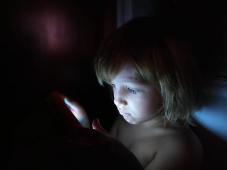 Cute little girl plays with a tablet computer. A child of 7 years old with a serious face and blond hair looks into a gadget. Copy space. Girl in the room. Dark and light wall. At home in the night.