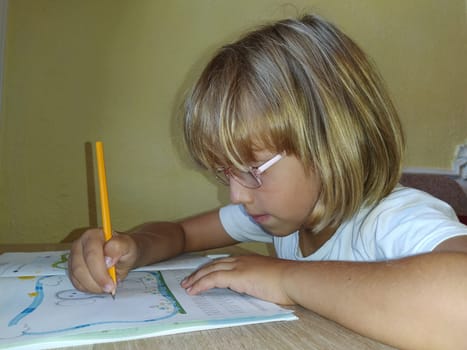 A 6-7 year old girl is reading a book or textbook and smiling. The child has blond hair, dark skin and glasses on his face. The child sits at the desk. yellow wall in the background.