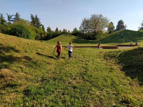 Sremska Mitrovica, Serbia - April 24, 2020- Children run in the park. A boy in a white T-shirt and blue trousers. Girl in a red tank top and trousers. Green lawn or field. People walk in a city park.