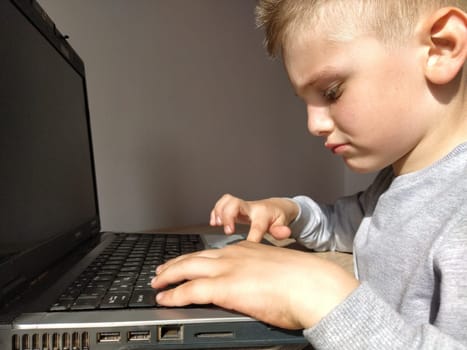 The small boy with the computer. The boy turns on the laptop. The black keyboard. A child with blond hair. Pensive face. Education concept. Gadget addiction concept.