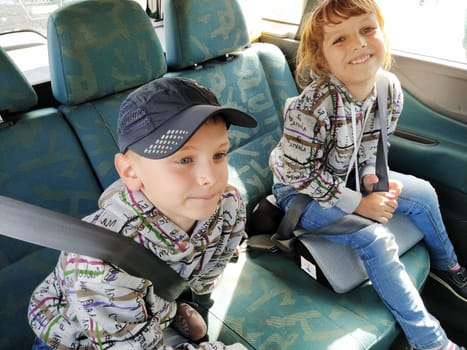 Children in the car. A boy and a girl are sitting in the back seat, laughing and grimacing. Children fasten their seat belts. A child in a cap. Children - twins of different faces dressed in the same clothes