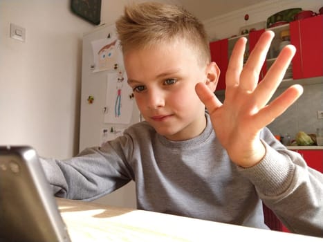 Inside shot of a blond child sitting at a table looking at the screen of a laptop or smartphone. Talk with mom or grandmother through a gadget. Online communication during self-isolation. Child waving.