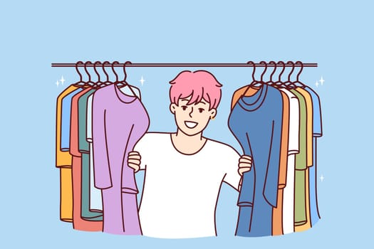 Young Cheerful man in t-shirt smiling peeking out from behind hanger for collection of clothes choosing new look. Guy with pink hair is standing in clothing store wanting to buy fashionable outfit