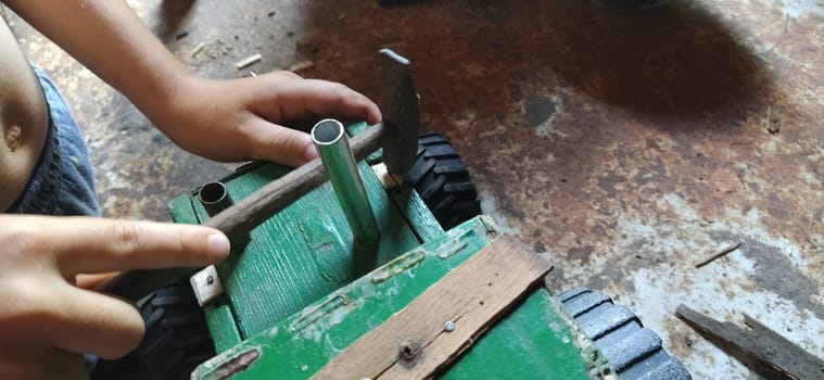 Hammer in hand. The boy is repairing a toy. Tools and people. Green wooden tractor with black plastic wheels. The child independently drives the nails into the surface. Skills of the master work.