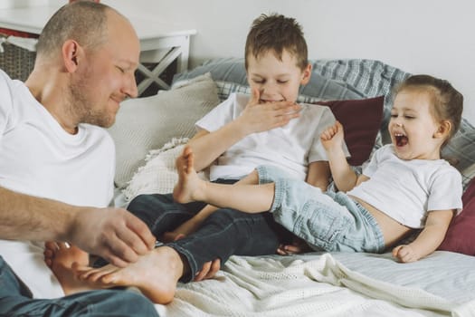 Father plays with two children on bed. Dad tickles kids feet. Family of daddy, girl and boy