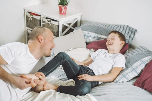 Father plays with his son 7-10 on bed. Dad tickles kids feet. Family having fun