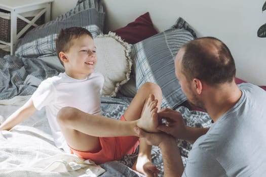 Father plays with his son 7-10 on bed. Dad tickles kids feet. Family having fun