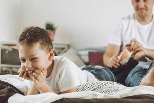 Father plays with his son 7-10 on bed. Dad tickles kids feet. Family having fun