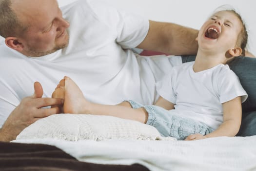 Father plays with little daughter 2-4 on bed. Dad tickles kids feet. Family having fun