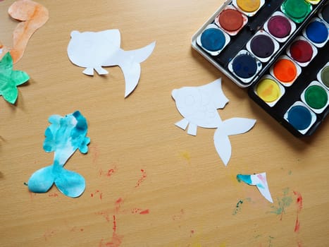 Desktop with drawing tools. A set of watercolor paints. Paper-cut, painted fish figurines. View from above. Lesson or creative task.
