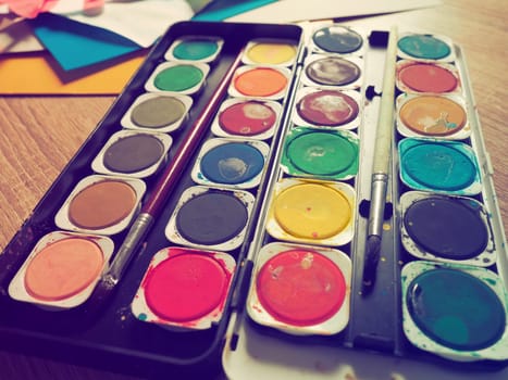 A set of watercolors of different colors for drawing. Multicolored round containers with bright paint. Tools for the lesson of drawing and labor. Colored paper. Red, blue, yellow, green colors