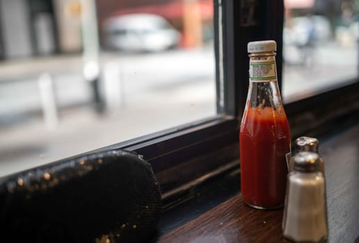 Defocused window looks out on a daytime city street. Ketchup bottle and salt shaker in foreground with black sequined evening bag purse. Dark wood window frames and table. No people, with excellent copy space.