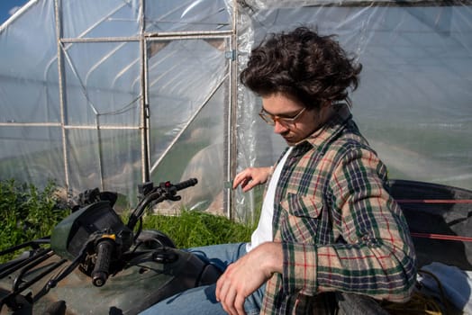 Young caucasion male farmer on an off road farm vehicle contemplating beside an organic vegetable garden greenhouse in natural golden hour sunlight with copy space.