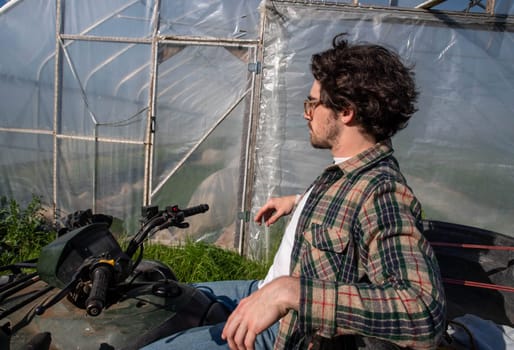 Young caucasion male farmer on an off road farm vehicle contemplating beside an organic vegetable garden greenhouse in natural golden hour sunlight with copy space.