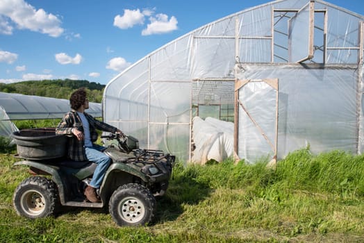 Young caucasian male farmer drives an off road vehicle to inspect a vegetable garden greenhouse on an organic farm. Farmer looks back at camera. Greenhouse door is open with covered rows of vegetables visible inside. Green grass and blue sky, shot in natural light with copy space.