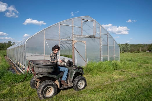 Young caucasian male farmer drives an off road vehicle by a vegetable garden greenhouse on an organic farm. Farmer looks back at camera. Greenhouse door is open with covered rows of vegetables visible inside. Green grass and blue sky, shot in natural light with copy space.