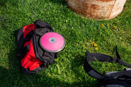 A 1 kg round pink track and field discus in a red athletic equipment bag lays off center in the grass. Shot in natural sunlight with no people and copy space.