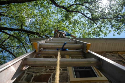 Low angle view of man on ladder cleaning gutters of old stone home with large leafy branches overhead, blue sky and clouds. Architectural features visible and one window.