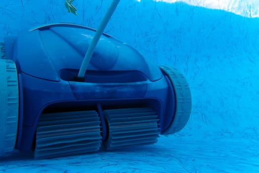 Residential blue vinyl pool liner being cleaned by a motorized wheeled vaccum cleaner with a single leaf in the water. Front wheels and beaters are visible and floating electric cord extends upward. Natural underwater light, no people, with copy space.