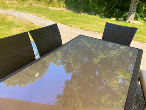 Beautiful outdoor summer table with glass top reflecting the green leaves on the tall trees above and blue sky in a green grass yard with cememt walkway. Three chairs are visible with no people, and copy space. Shot in natural light.