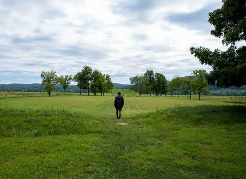 A young man walks toward the Native American Hopewell Culture prehistoric Seip Earthworks burial mound in Ohio. Ancient large long mound. Grass is neatly trimmed with trees and dramatic sky. Copy space