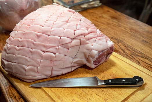 Antique wooden table with wood grain texture and cutting board with black-handled chef's knife and a big skin on bone-in pork shoulder picnic roast. Skin has been cut into diamond cross hatch pattern. No people with copy space