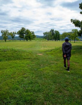 A young man walks toward the Native American Hopewell Culture prehistoric Seip Earthworks burial mound in Ohio. Ancient large long mound. Grass is neatly trimmed with trees and dramatic sky. Copy space