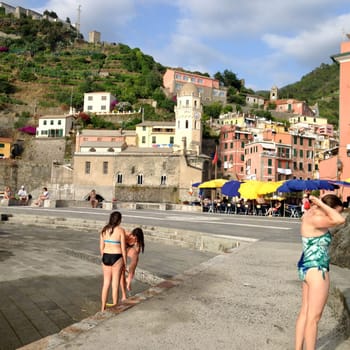 08-05-2014, Vernazza, Italy, Waterfront village scene with church and swimmers Beautiful marina with swimmers and outdoor restaurant with colorful table umbrellas in the foreground of this centuries old village in the Cinque Terre region of Italy. Santa Margherita di Antiochia Church with bell tower and clock as well as ancient houses and green hills on a summer day.