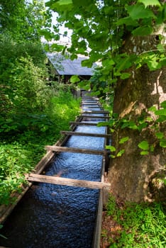 A wooden colonial American sluice leads away from the camera next to a large tree into a building in the background. Green nature setting with no people. Shot in natural sunlight with copy space.