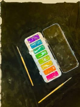An open pan of watercolor pains with two brushes on a black art background cloth over a table visible around two edges digital art illustration with copy space.
