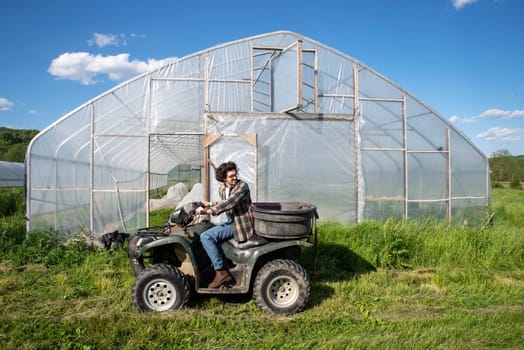 Young caucasian male farmer drives an atv by a vegetable garden greenhouse on an organic farm.Greenhouse door is open with covered rows of vegetables visible inside. Green grass and blue sky, shot in natural light with copy space.