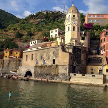 08-05-2014, Vernazza, Italy, Waterfront village scene with church and swimmers Beautiful marina with swimmers and boats in the foreground of this centuries old village in the Cinque Terre region of Italy. Santa Margherita di Antiochia Church with bell tower and clock as well as ancient houses and green hills on a summer day.