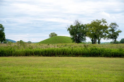 Grass covered prehistoric Hopewell culture burial mound at Siep Earthworks in Ohio under a dramatic cloudy sky with green grass and meadow foreground.